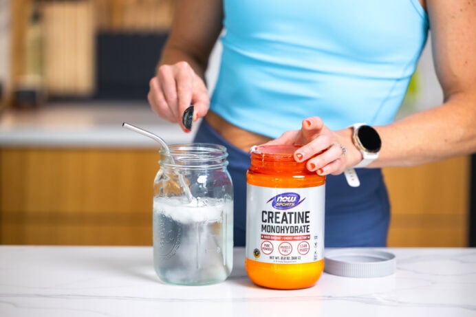 Women pouring a tsp scoop of creatine monohydrate into a glass cup.